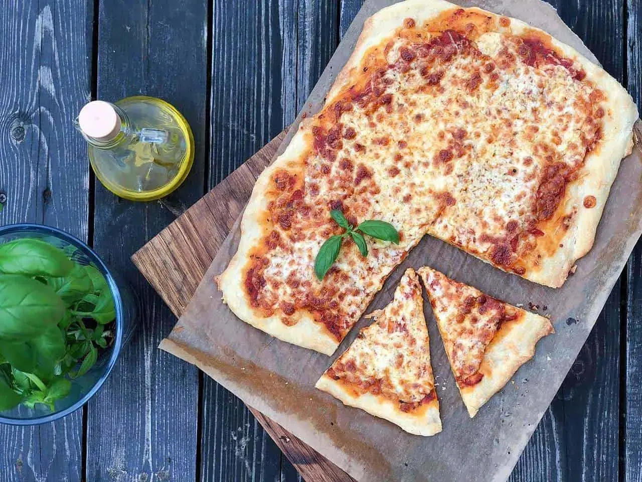 Pyszna pizza margherita z roztopionym serem i świeżą bazylią, obok butelka oliwy i miseczka ziół.
