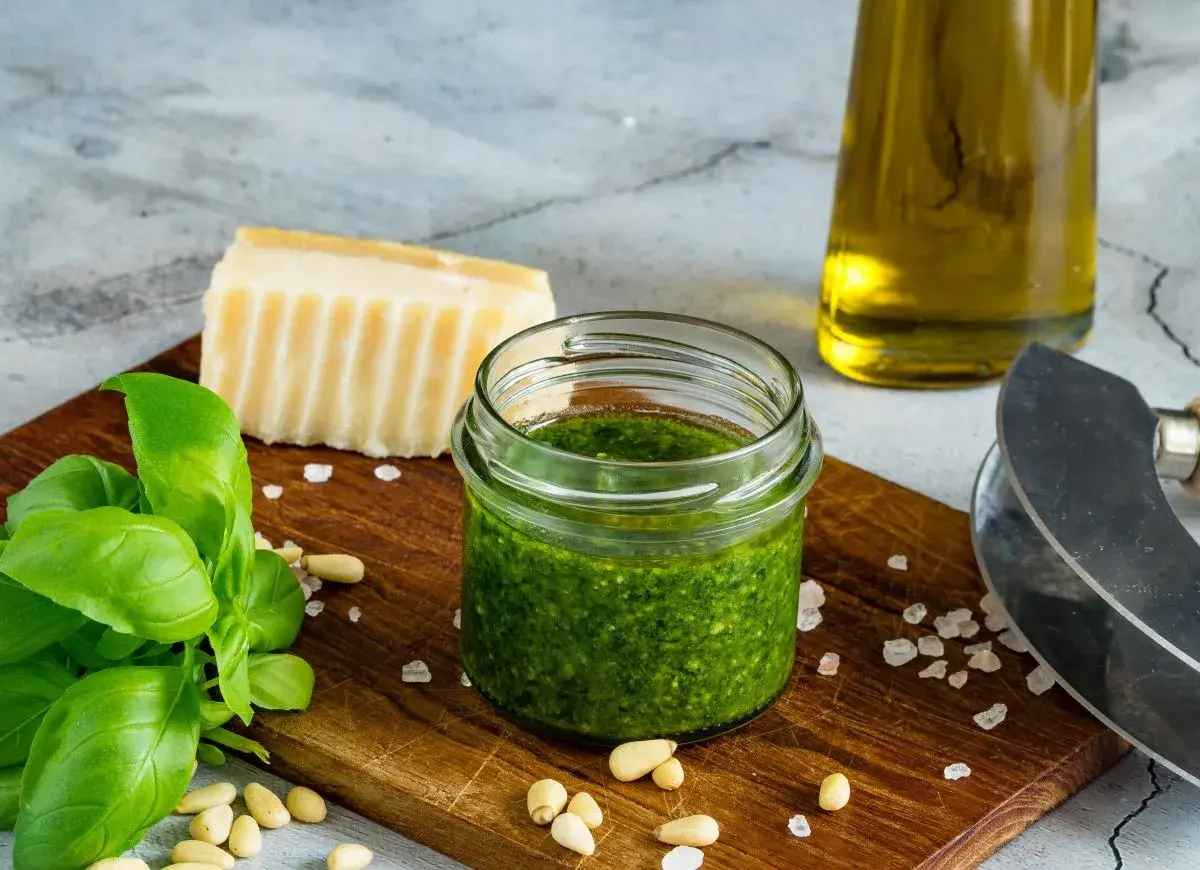 Słoiczek domowego pesto z bazylii, obok świeże liście bazylii, orzeszki piniowe, kawałek parmezanu i oliwa.