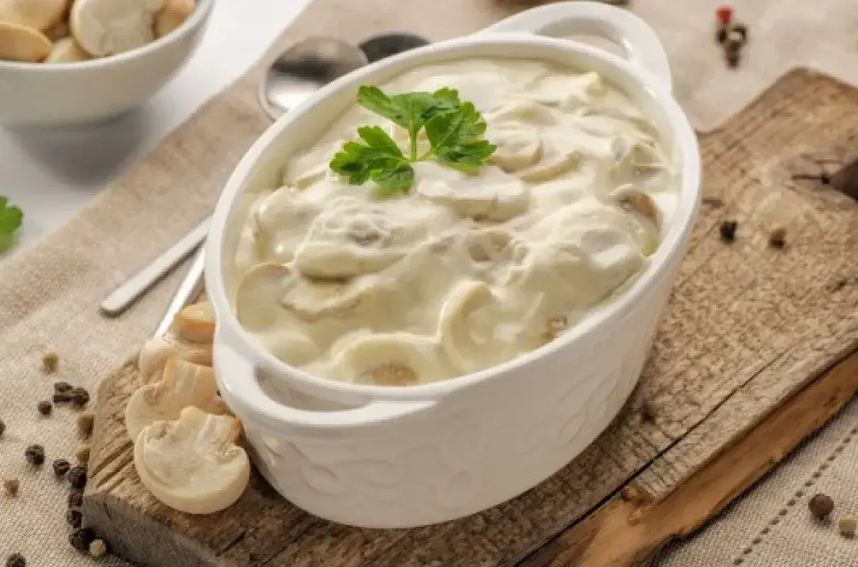 Aksamitny sos pieczarkowy w białej miseczce, udekorowany natką pietruszki. Obok świeże pieczarki i ziarna pieprzu.