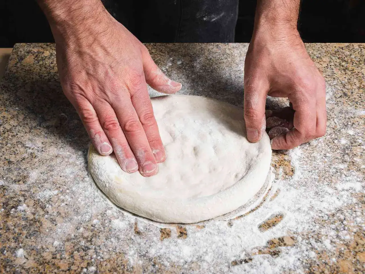 Dłonie formują ciasto na pizzę na grubym cieście, posypane mąką na blacie.