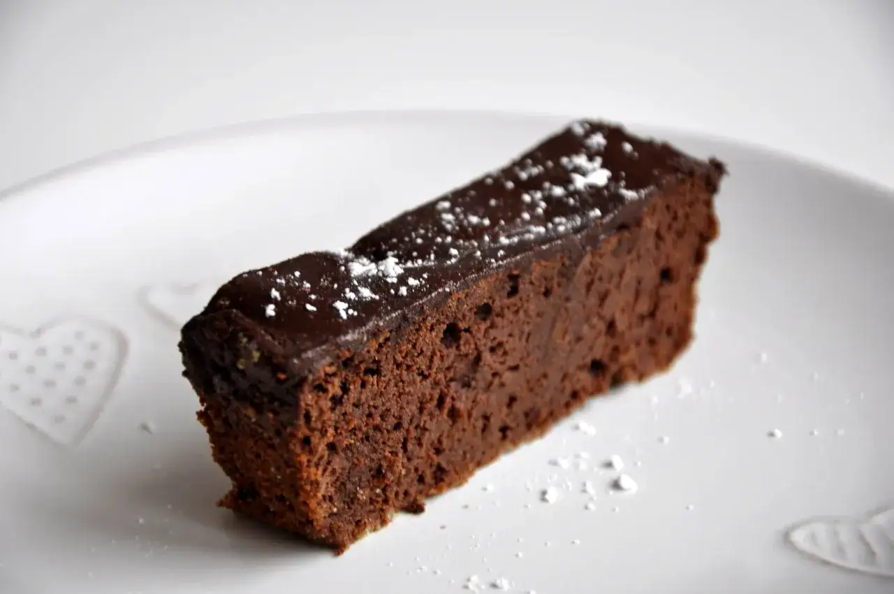 Czekoladowy brownie z fasoli, polany błyszczącą polewą i posypany cukrem pudrem, kusi na białym talerzu.
