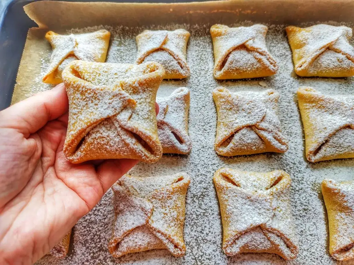 Ręka trzyma jedno z wielu pysznych ciastek z jabłkami, posypanych cukrem pudrem.