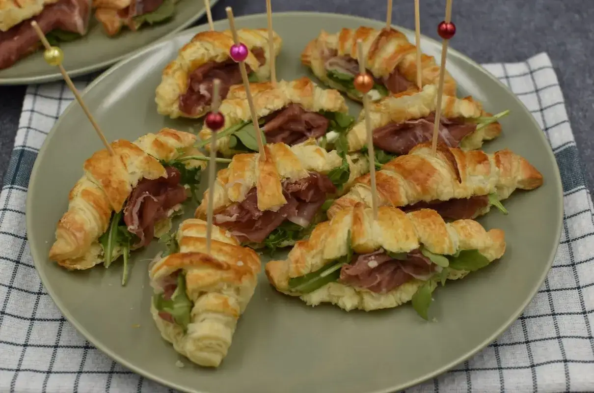 Mini rogaliki z ciasta francuskiego z szynką parmeńską i rukolą, ozdobione kolorowymi patyczkami. Idealne na przyjęcie.