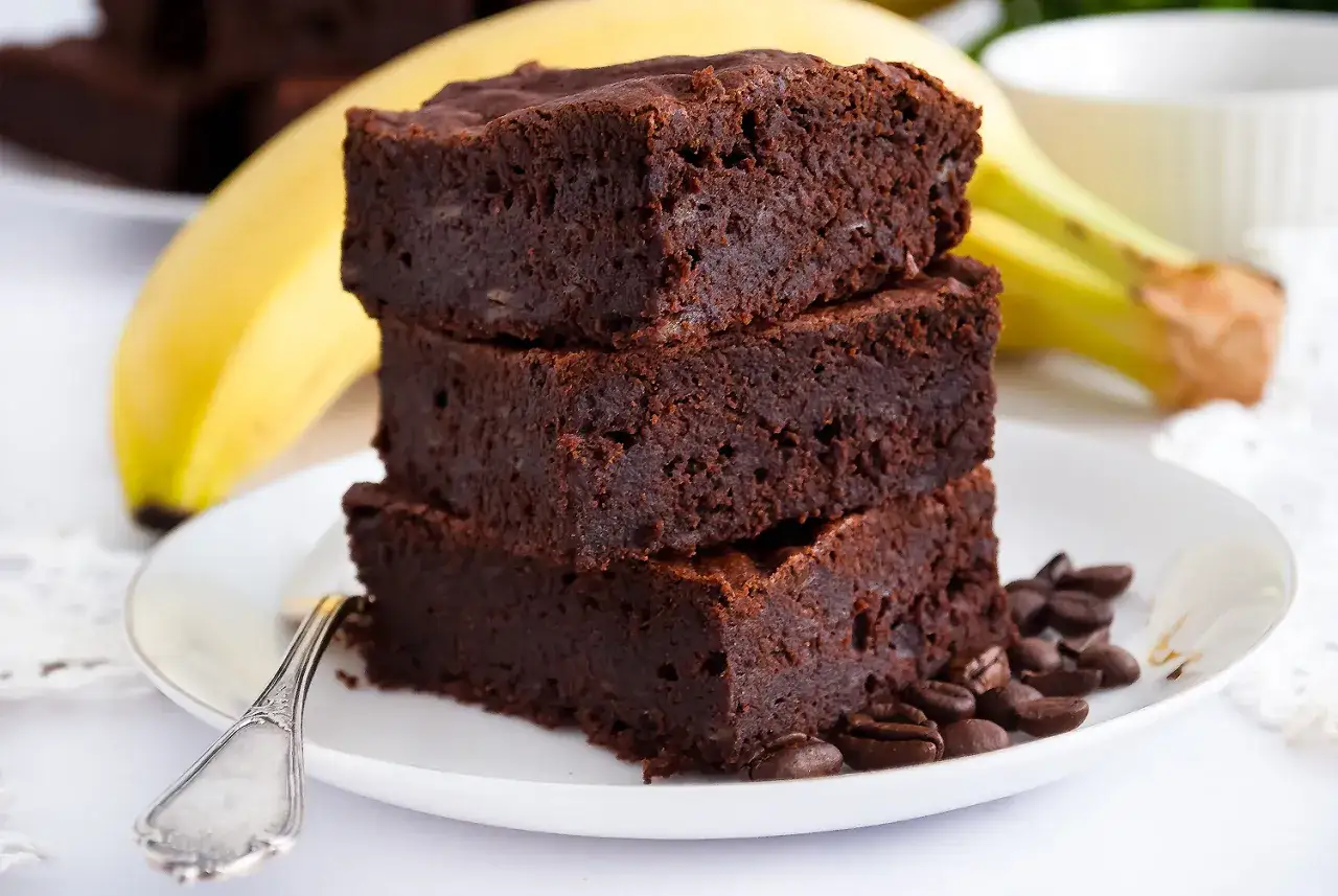 Stos puszystych brownie z bananami, ozdobiony ziarnami kawy, na talerzu z bananem w tle.