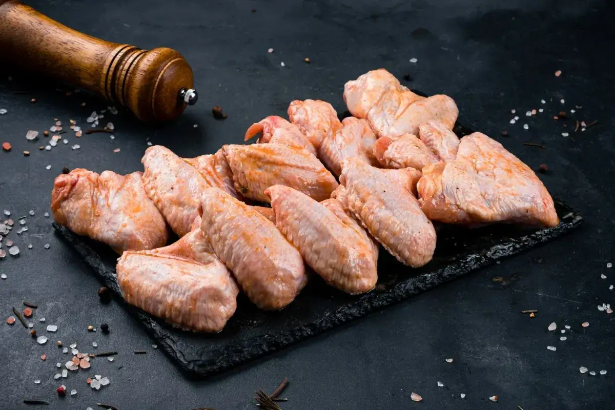Surowe skrzydełka kurczaka, przygotowane do pieczenia, zanurzone w aromatycznej solance do mięsa. Obok młynek do pieprzu i gruboziarnista sól.