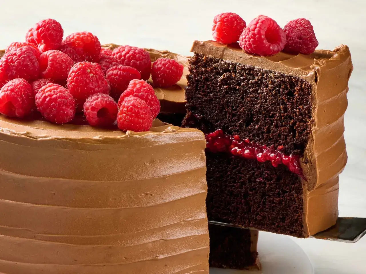 Pyszny brownie z malinami, z czekoladowym kremem i świeżymi malinami na wierzchu. Kawałek ciasta jest krojony łopatką.