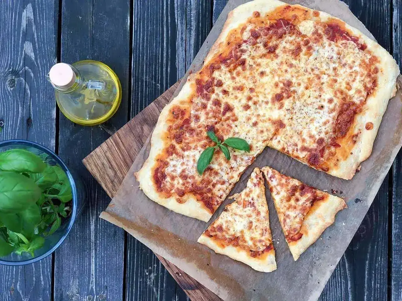 Pyszna pizza margherita z ciągnącym się serem i świeżą bazylią, obok buteleczka oliwy i miseczka z ziołami.