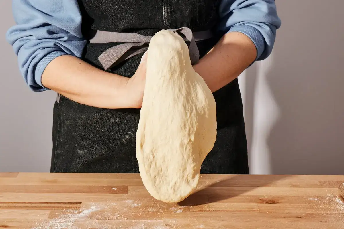 Ręce formują elastyczne ciasto na pizzę, które wisi swobodnie, gotowe do dalszej obróbki.