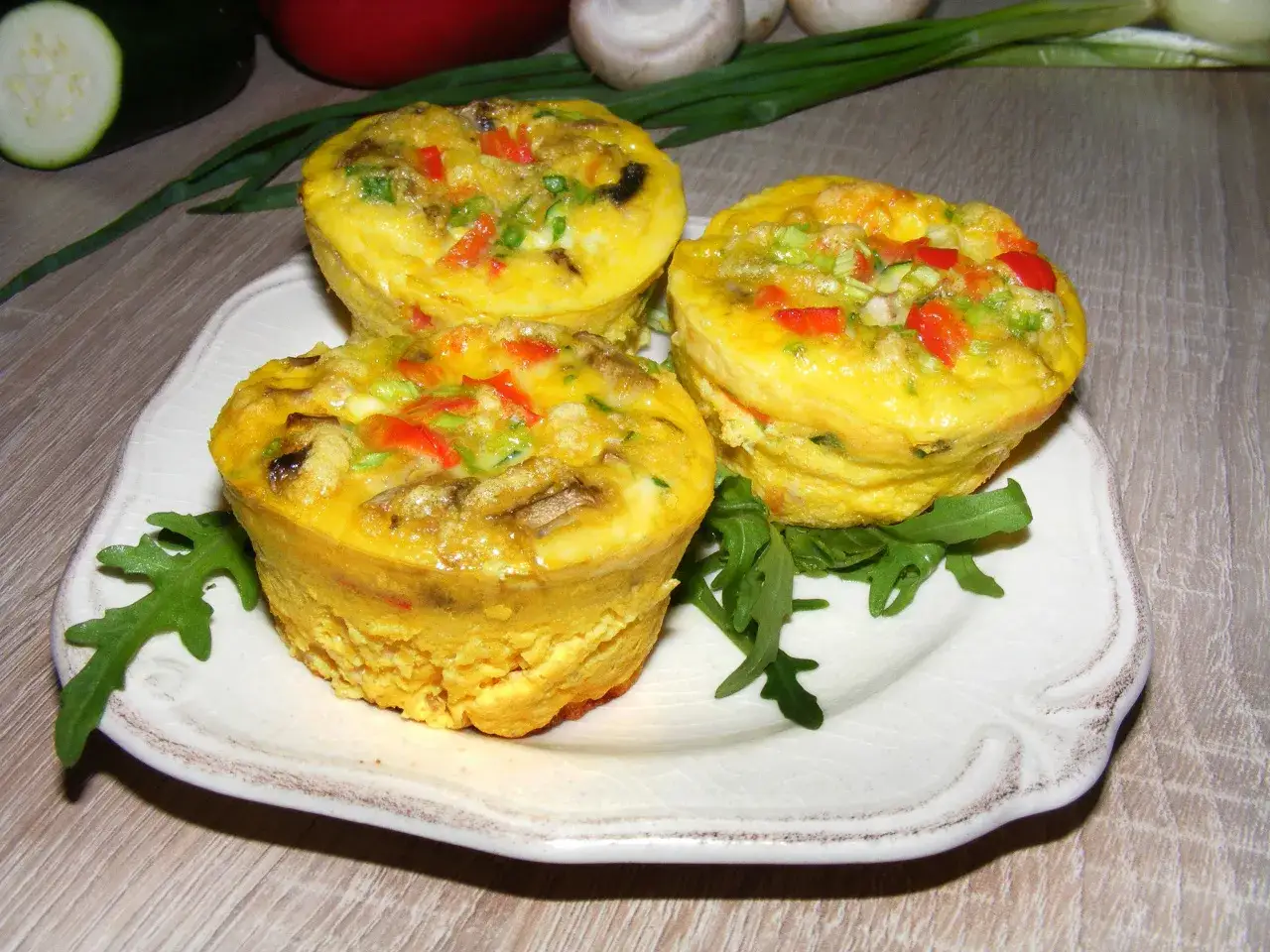 Trzy apetyczne muffiny jajeczne z warzywami, podane na talerzu z rukolą.