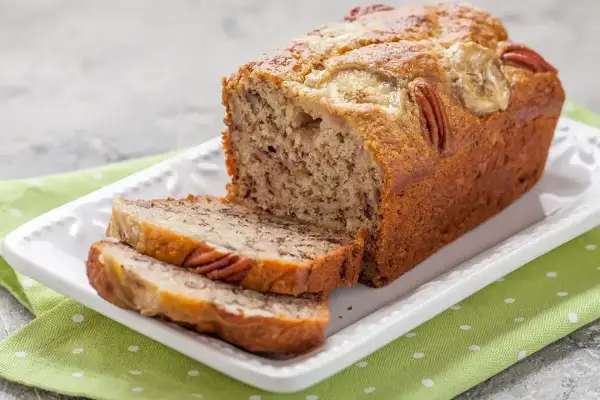Pyszny chlebek bananowy z orzechami pekan i plasterkami banana na wierzchu, pokrojony na kromki, na białej paterze.