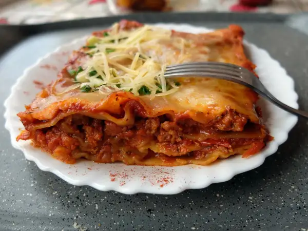 Pyszna lasagne bolognese z warstwami makaronu, mięsa mielonego i sosu pomidorowego, posypana serem i natką pietruszki. Widelec czeka na pierwszy kęs.