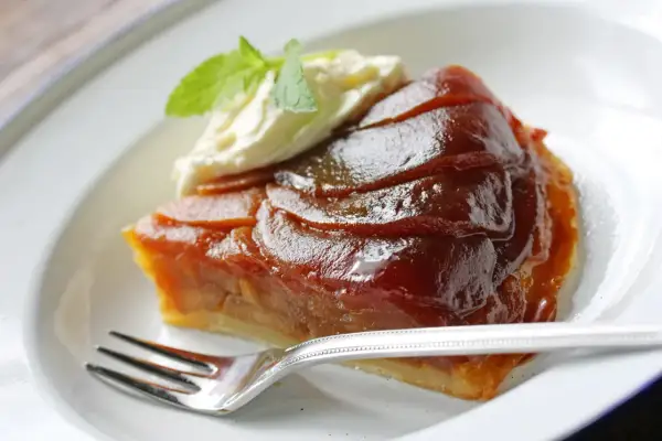 Kawałek karmelizowanej tarta tatin z jabłkami, podany z bitą śmietaną i miętą, na białym talerzu z widelcem.