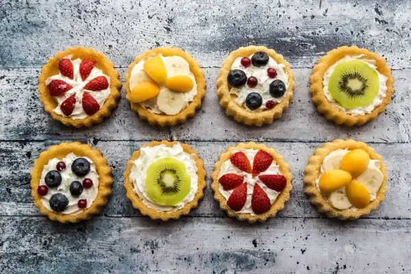 Osiem kolorowych tartaletek z owocami: truskawkami, borówkami, kiwi, bananami i morelami, na kremowym nadzieniu.