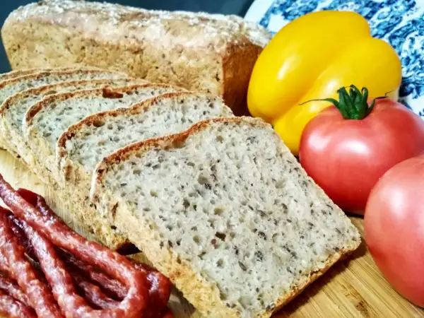 Pyszny chleb nocny na zakwasie, pokrojony w kromki, obok wędzonej kiełbasy, pomidorów i żółtej papryki.
