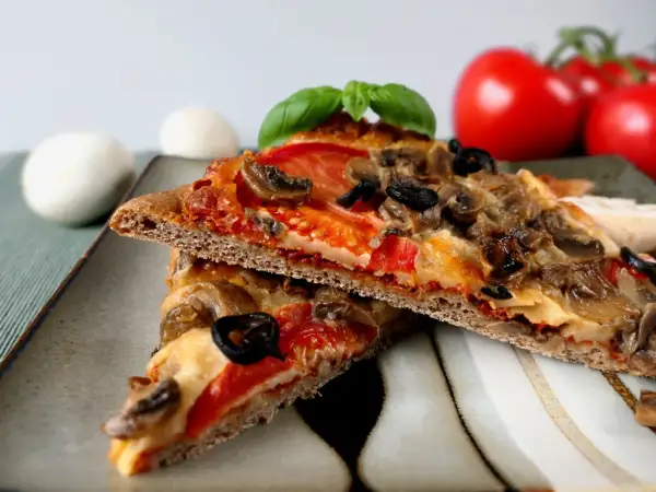Dwie kromki pizzy na cieście z mąki pełnoziarnistej z pieczarkami, oliwkami i pomidorami.