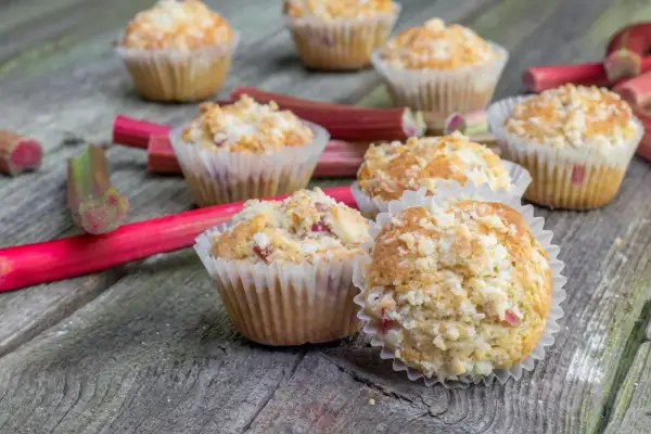 Pyszne muffinki z rabarbarem, posypane kruszonką, leżą na drewnianym blacie obok świeżych łodyg rabarbaru.