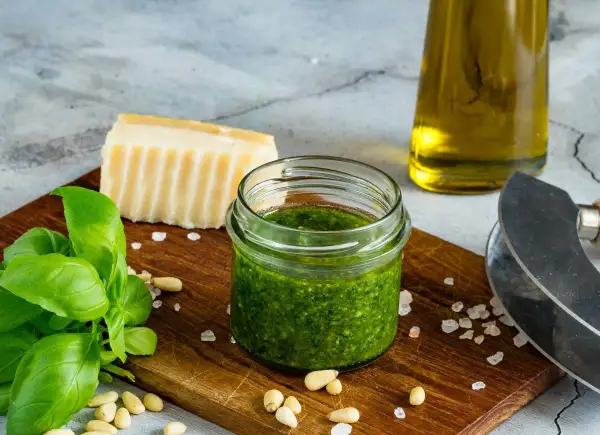 Słoiczek domowego pesto z bazylii, obok świeże liście bazylii, orzeszki piniowe, kawałek parmezanu i oliwa.