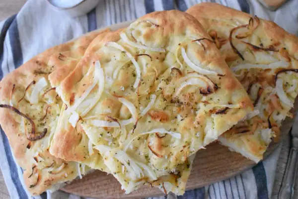 Puszysta focaccia z cebulą, posypana ziołami i solą, prezentuje się apetycznie na drewnianej desce.
