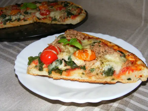 Kawałek pysznej pizzy z tuńczykiem, pomidorkami i bazylią na talerzyku. W tle cała pizza.